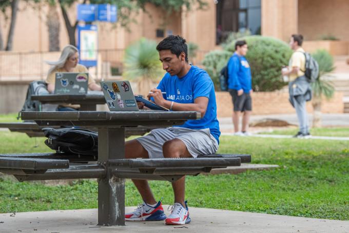 Student studying on his laptop at a table outside with another student studying and two students talking in the background.