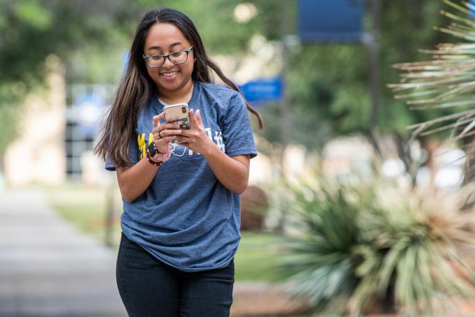 Student walking and looking at her phone while smiling.